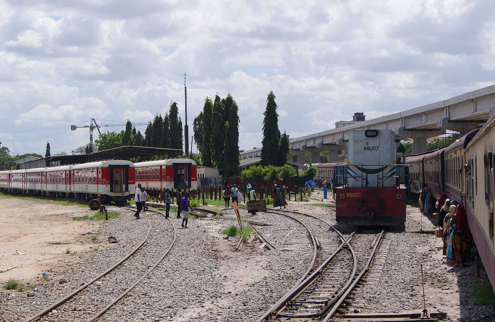 Daressalam Kamata Station im Oktober 2019: Am zur Abfahrt bereitgestellten Zug rangiert die Lok 88U07 vorbei. Links stehen moderne Reisezugwagen aus China, rechts oben ist die normalspurige Hochbahn zu sehen.