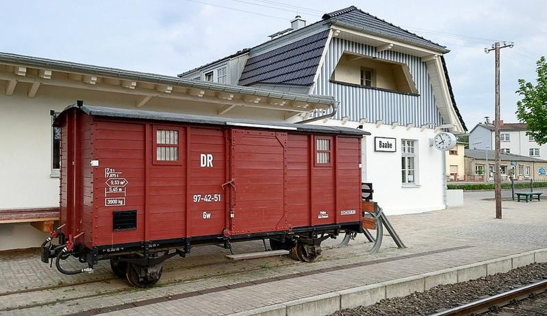 Vor dem im alten Stil neu errichteten Empfangsgebäude von Baabe dient der Güterwagen 97-42-51 derzeit als Lagerraum.