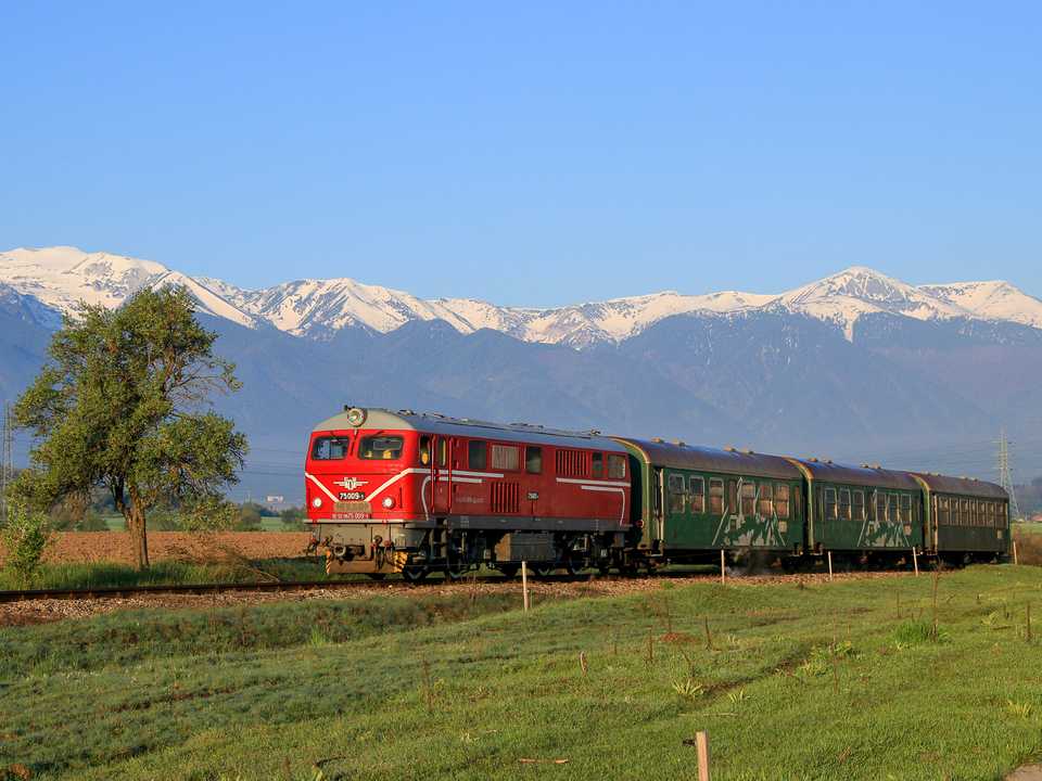 Am Morgen des 12. Mai 2022 verlässt die Diesellok 75.009 mit den Wagen des Personenzuges 16102 den Kreuzungsbahnhof Razlog. Im Hintergrund erhebt sich das Pirin-Gebirge.
